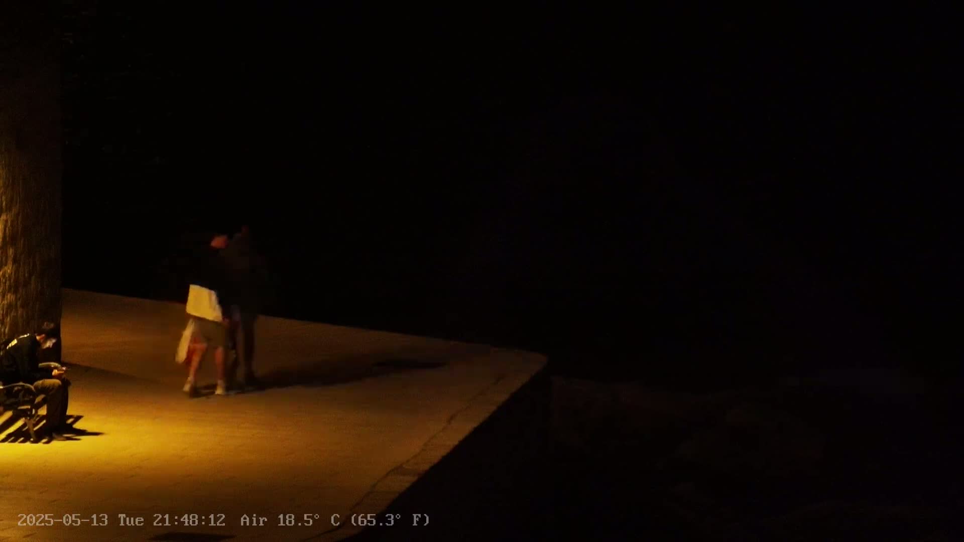 At night, under a dark sky with an air temperature of 18.5°C, two people stand close together on a dimly lit, paved surface near a dark wall, while a person sits alone on a bench.