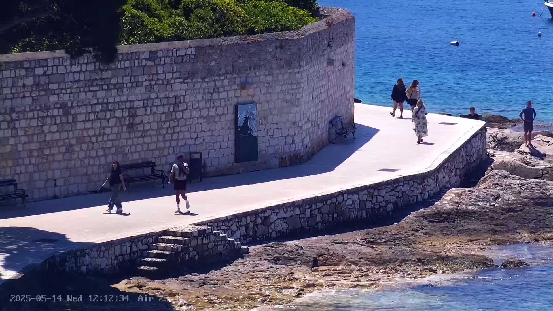 Several people stroll along a stone walkway next to a sea wall overlooking a clear blue sea on a sunny day.