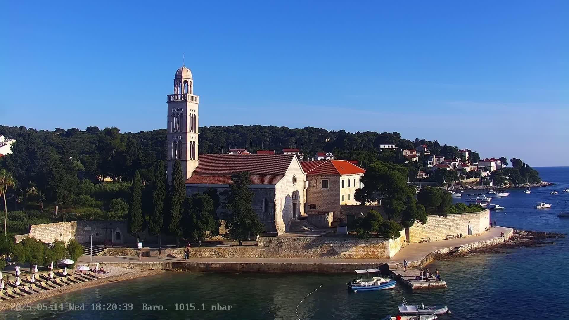 An aerial view shows a church with a tall bell tower situated on a coastal area with a clear blue sky, several boats in the water, and a beach with lounge chairs.