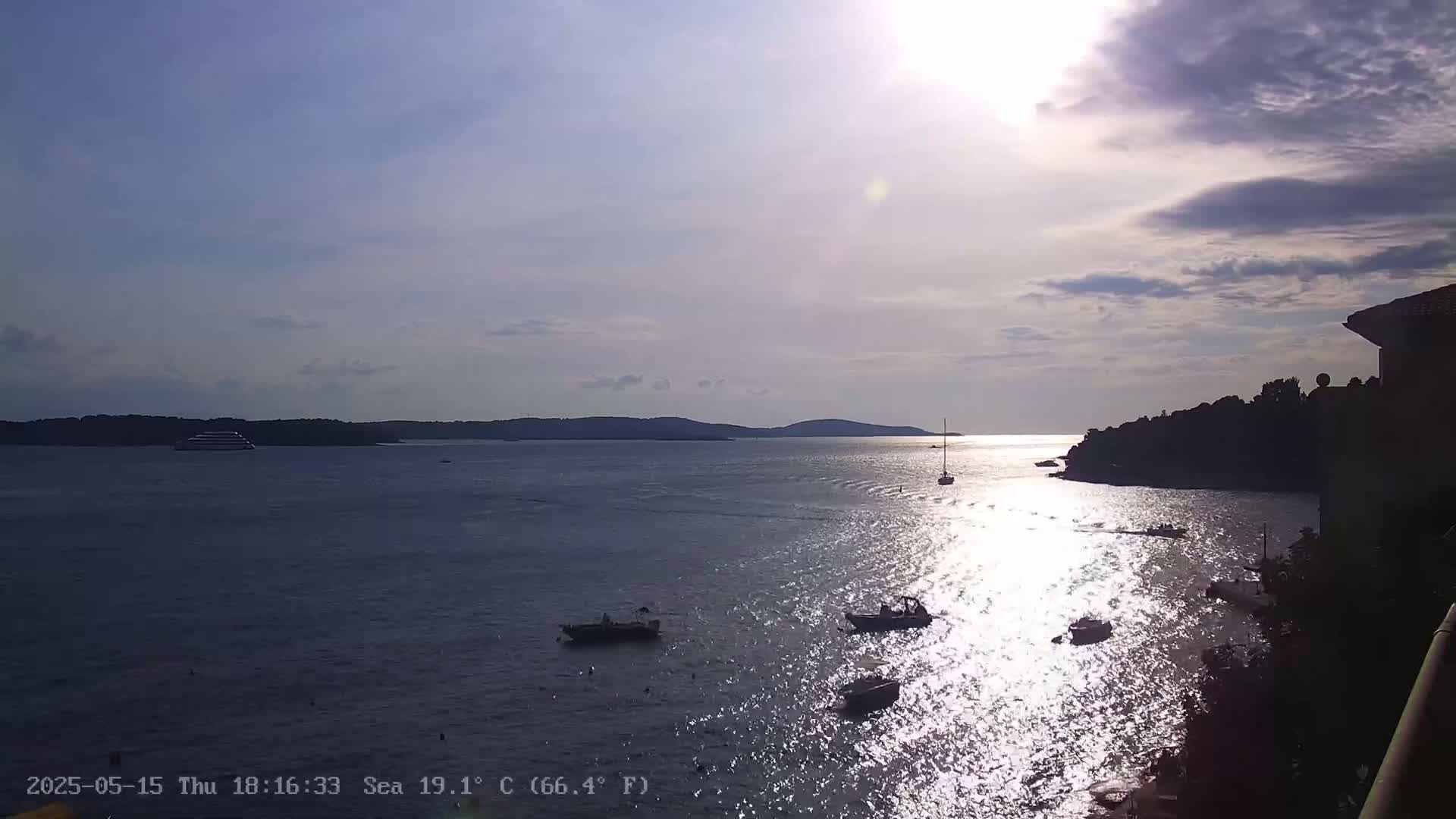 A calm, sunlit sea with several small boats and a larger vessel in the distance, under a partly cloudy sky.