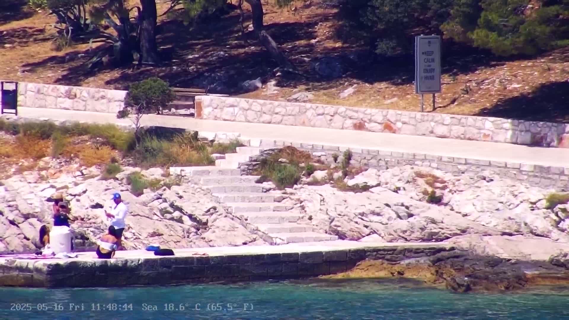 A sunny waterfront scene shows people relaxing near a stone staircase leading down to clear blue water.