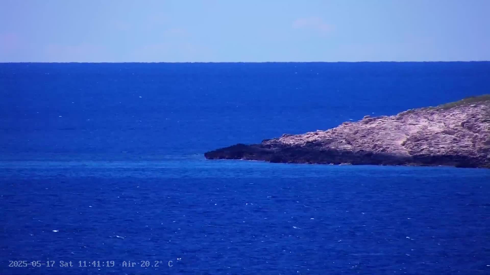 A rocky, light-pinkish shoreline juts into a deep blue sea under a clear sky.