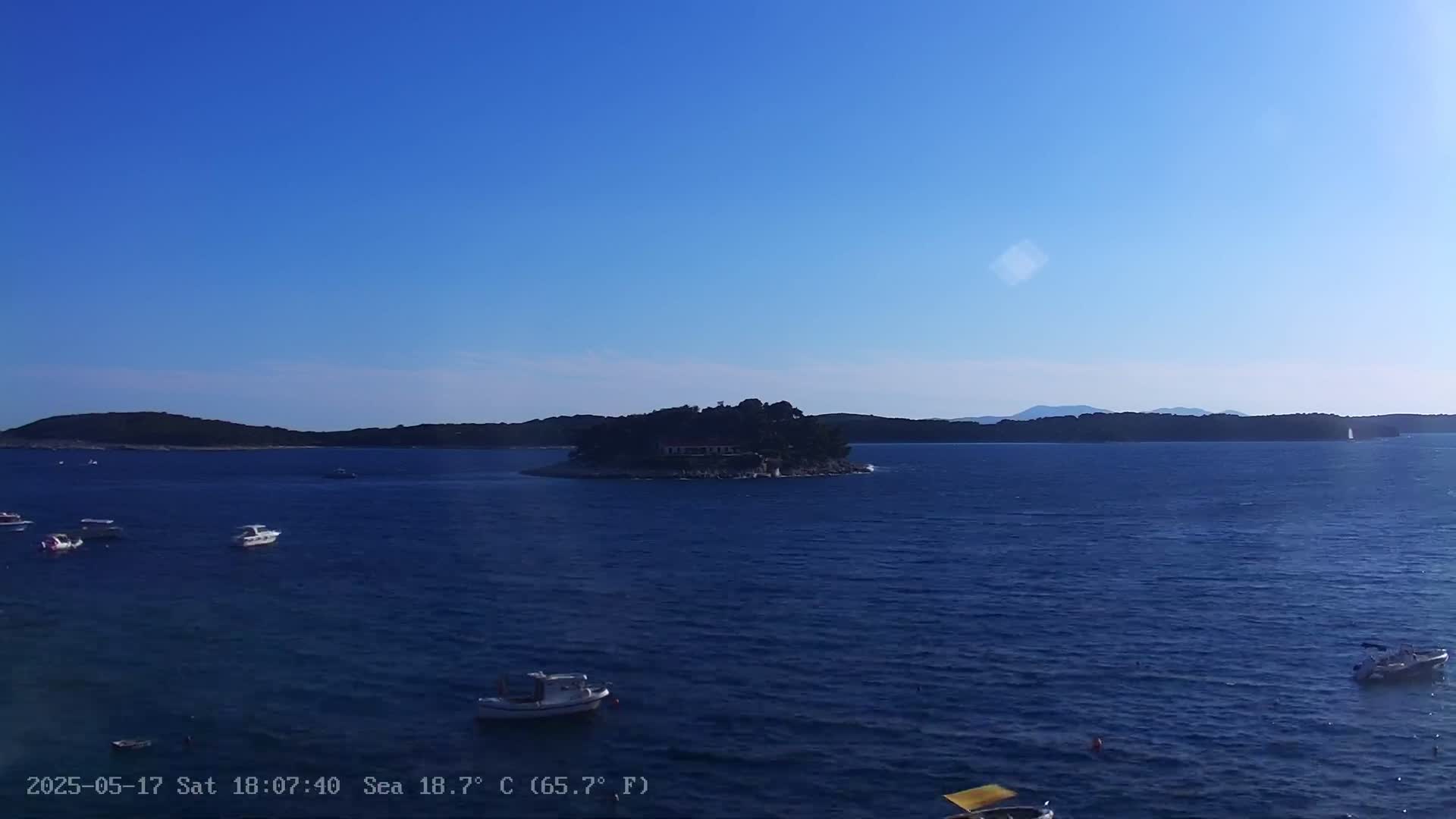 A small island with buildings sits in a calm, blue sea under a clear blue sky, with several small boats scattered in the water.