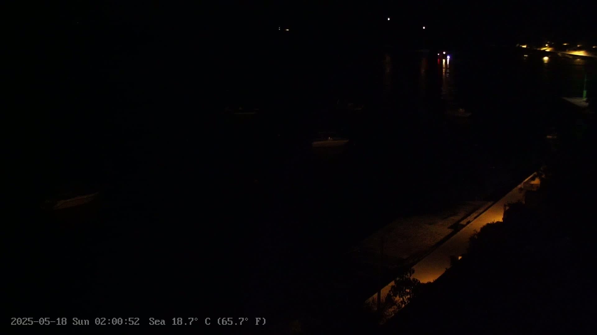 A nighttime, low-light view of a dark body of water with a dimly lit walkway along a shoreline, and a few faint lights visible in the distance.