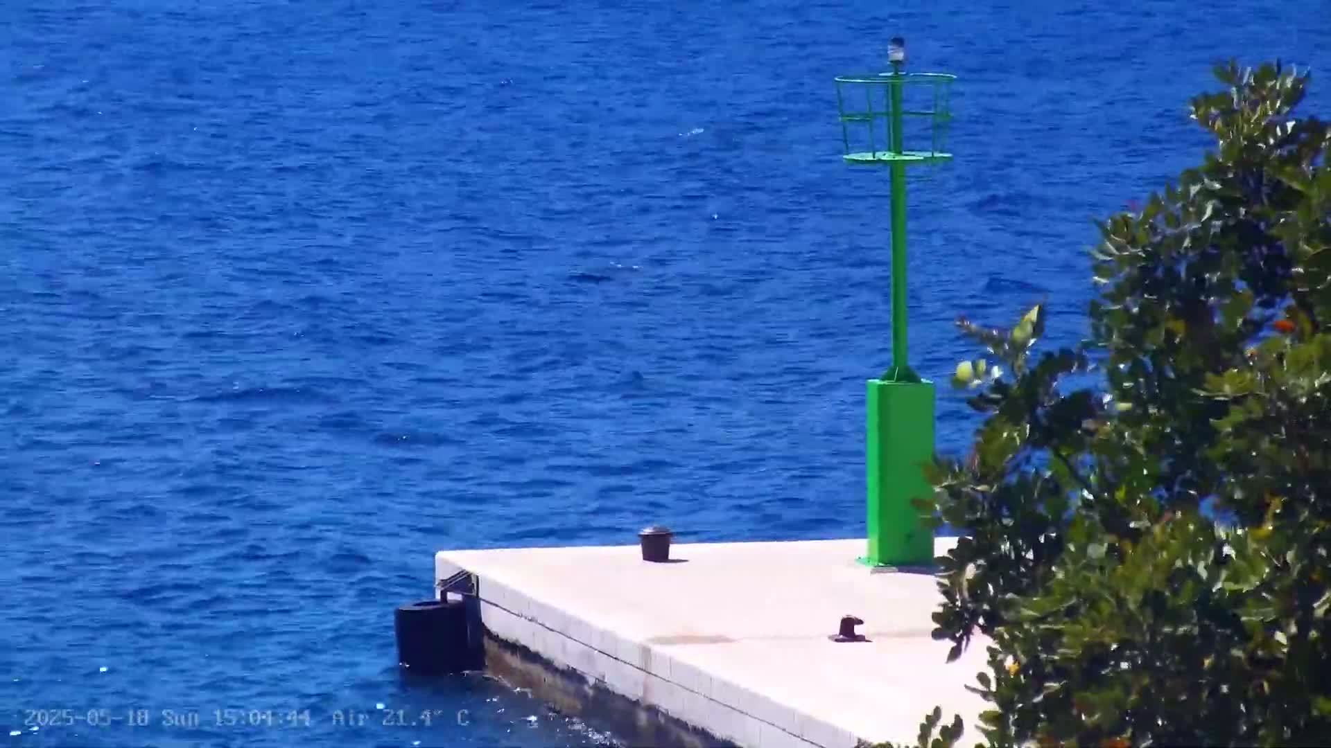A green navigational marker stands on a small, light-colored pier extending into a calm, deep blue sea under a sunny sky.