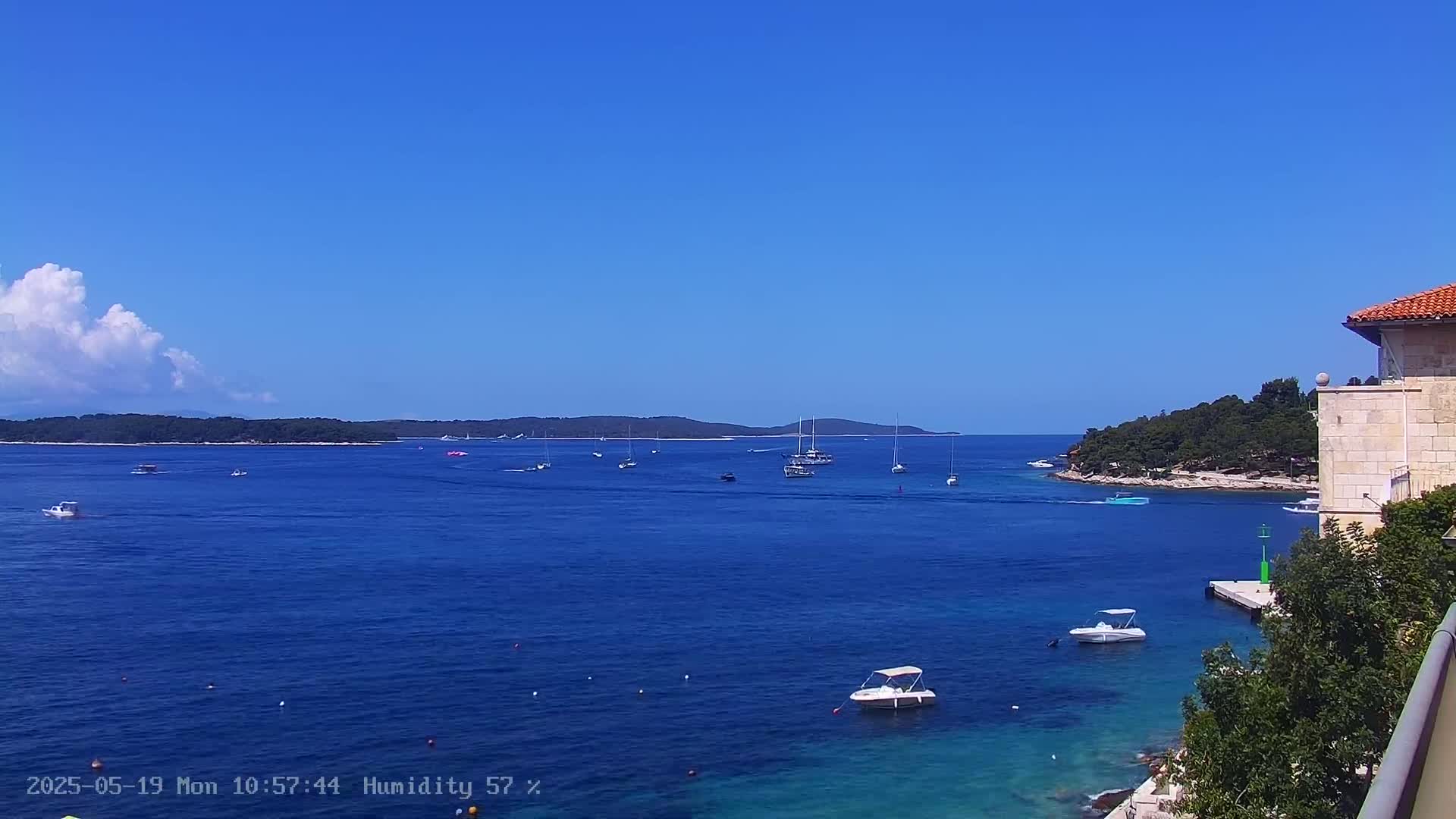 A clear blue sky overlooks a calm, blue sea dotted with several boats and small islands, partially visible from behind a stone building.