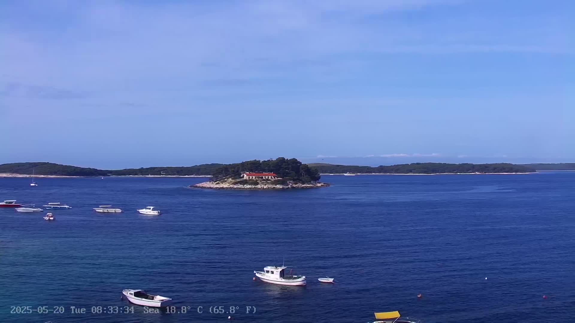 Several small boats are scattered across a calm, blue sea, with a small island featuring a building visible in the middle distance under a clear sky.