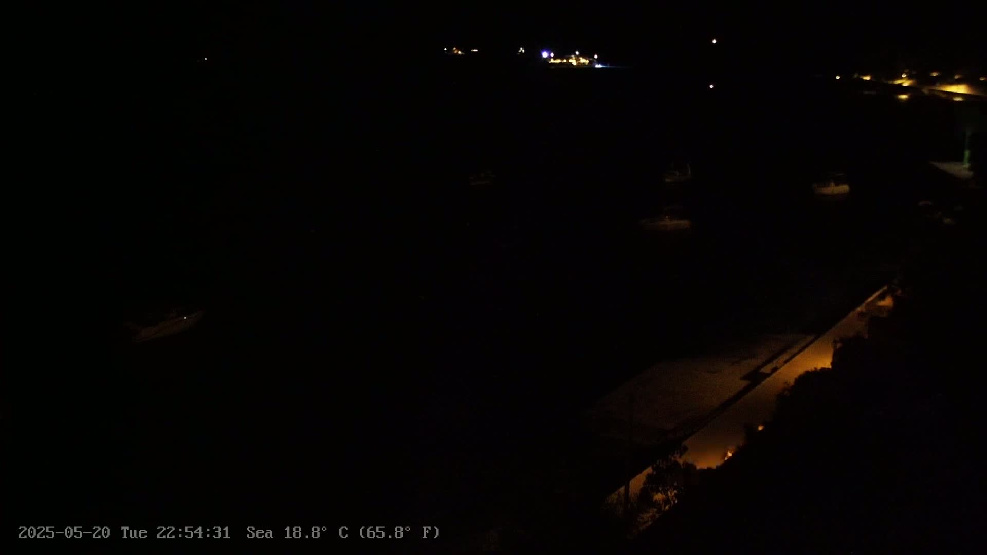 A dark nighttime scene shows a body of water with dimly lit boats and a shoreline with sparse, illuminated areas.