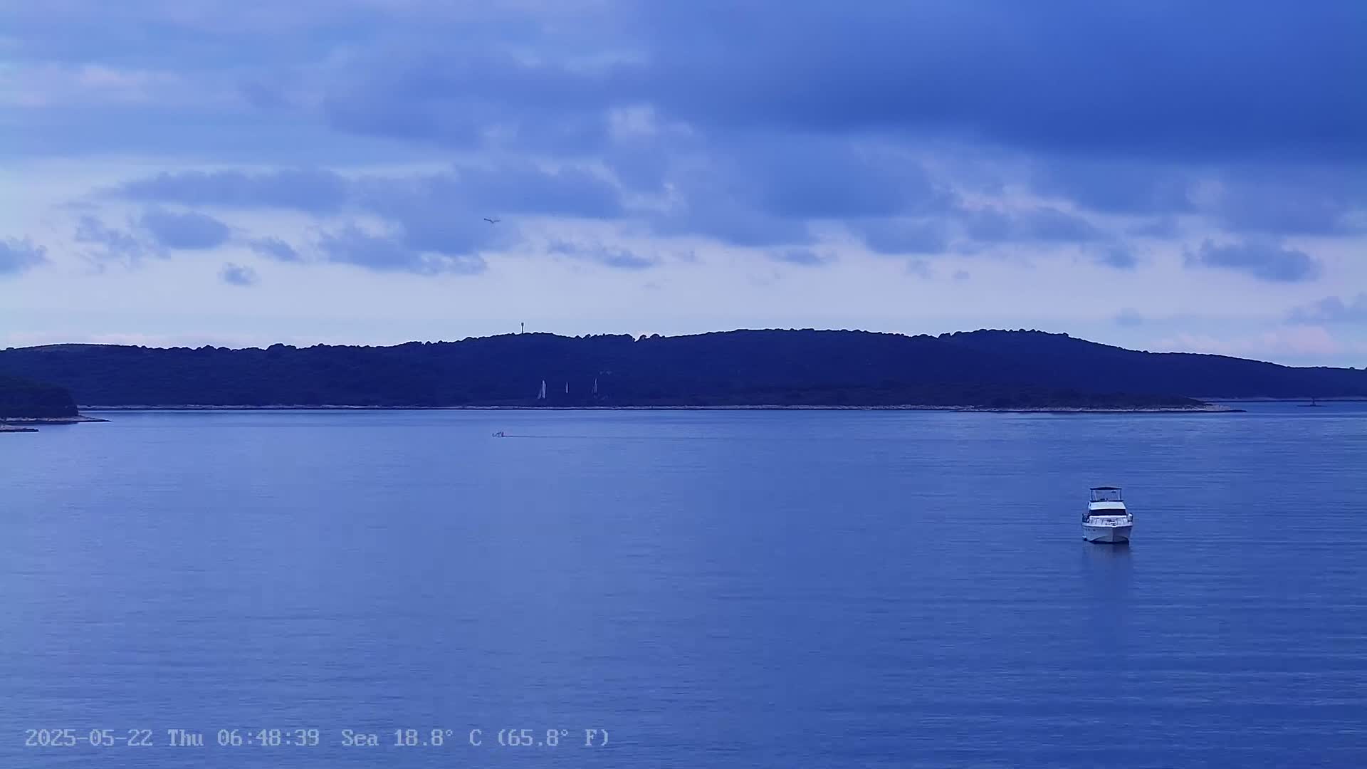 A single boat sits in calm blue water, with a dark, distant shoreline under a partly cloudy sky.