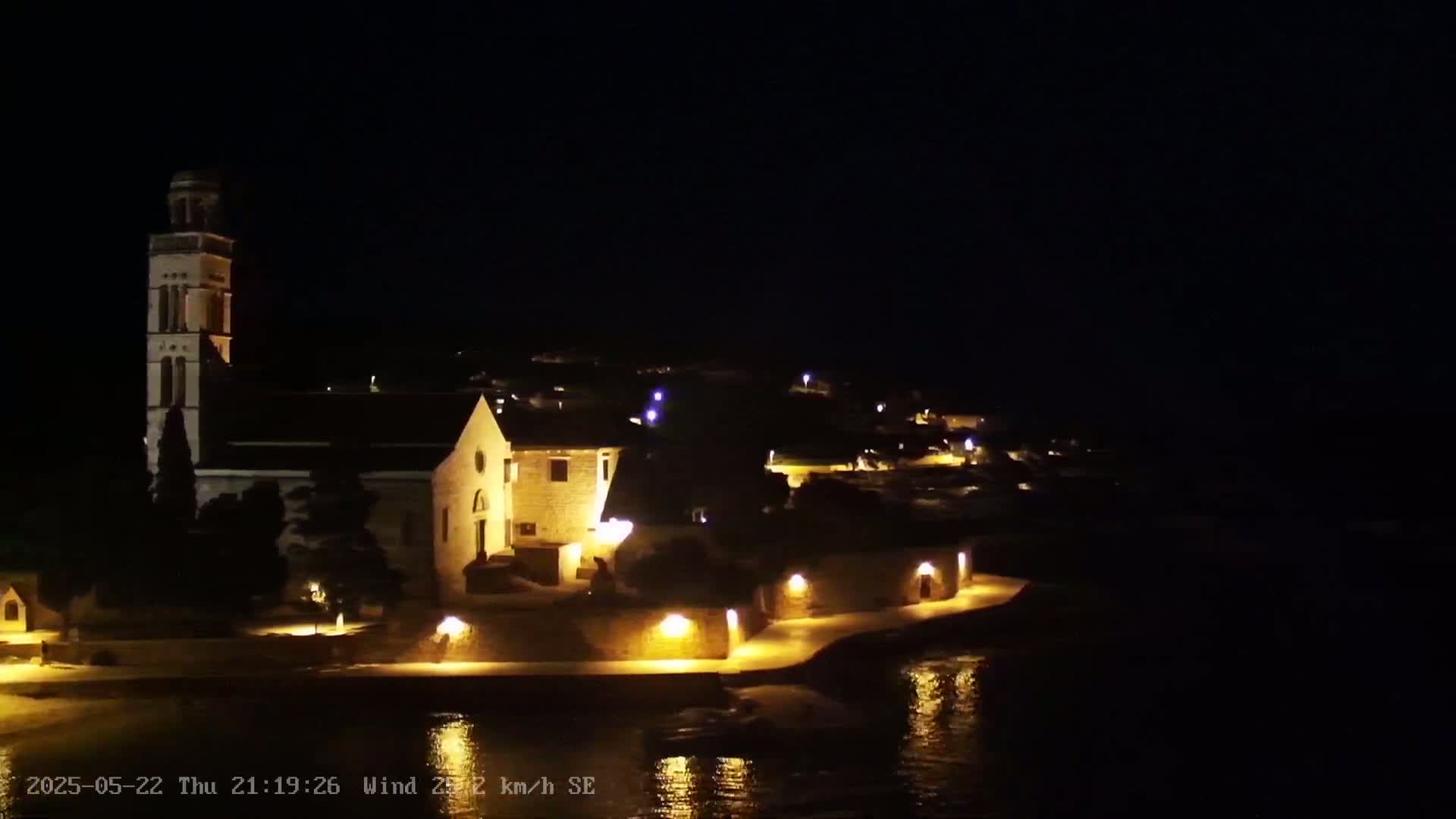 A church and village are illuminated at night next to a calm body of water under a dark sky.