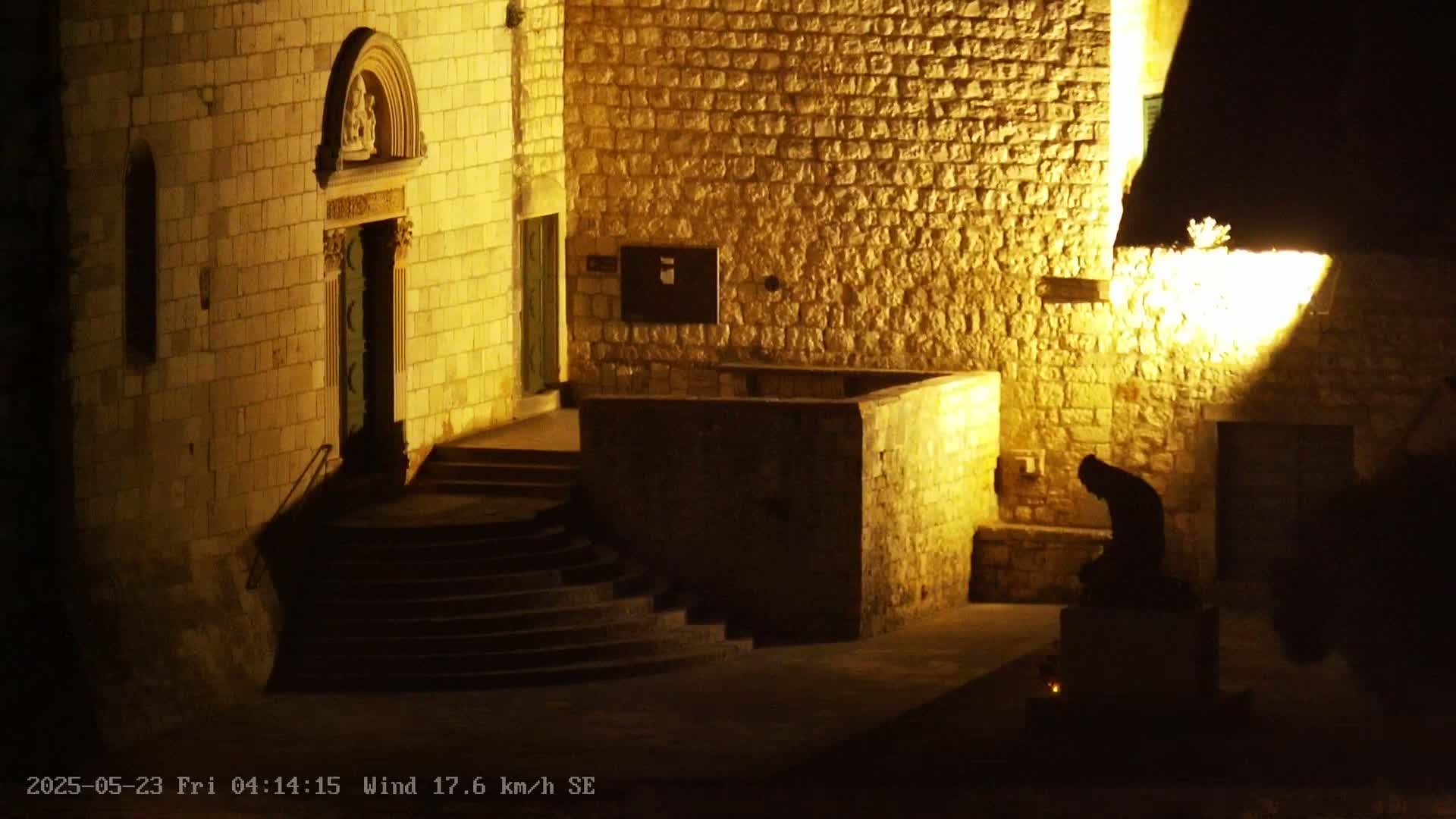 A stone building is illuminated at night by a light source, with a dark statue kneeling on a pedestal in the foreground.