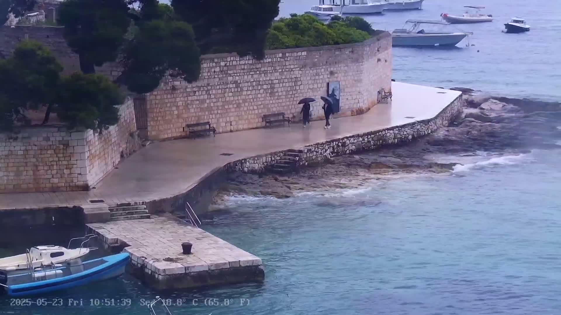 Two people with umbrellas walk along a stone walkway beside a calm sea during a rainy day, near several moored boats and a stone wall.