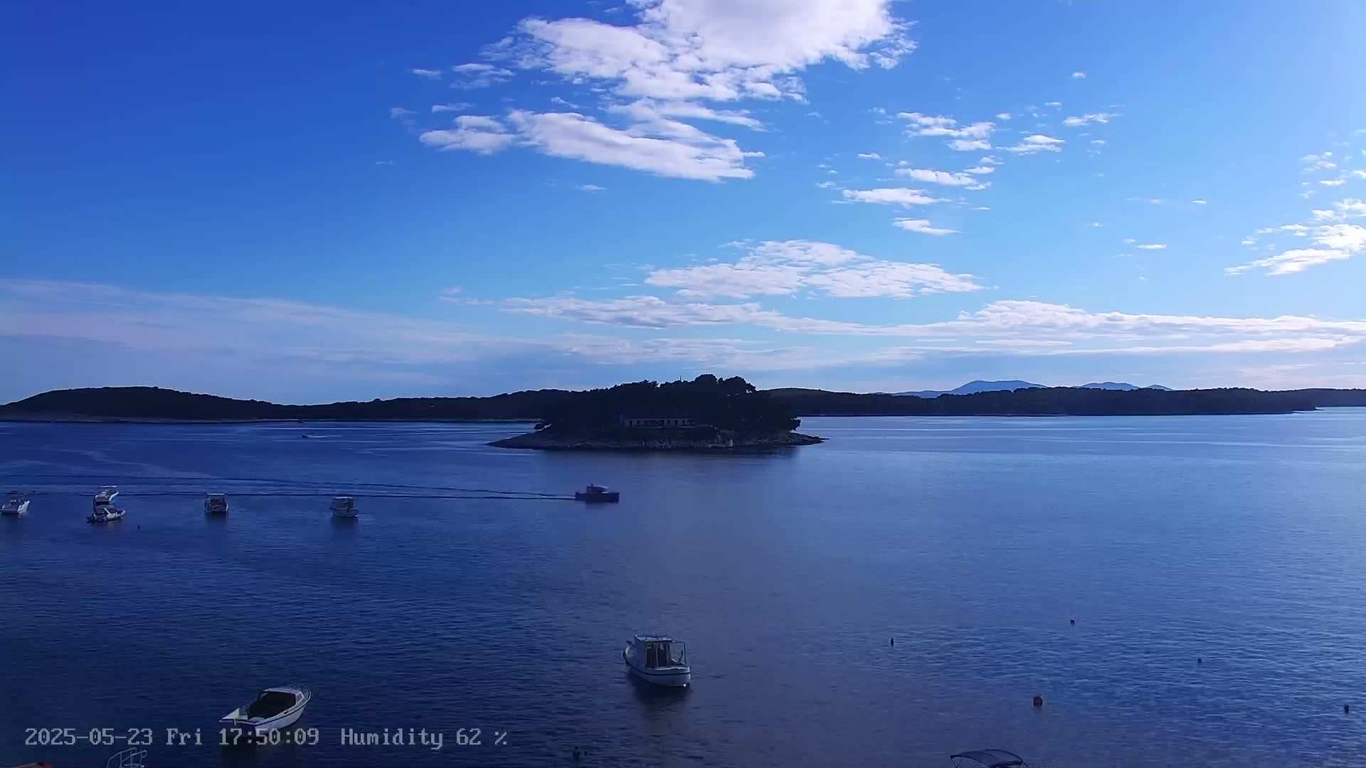 Several small boats are scattered across a calm, blue sea in front of a small, tree-covered island under a mostly clear, blue sky with scattered clouds.