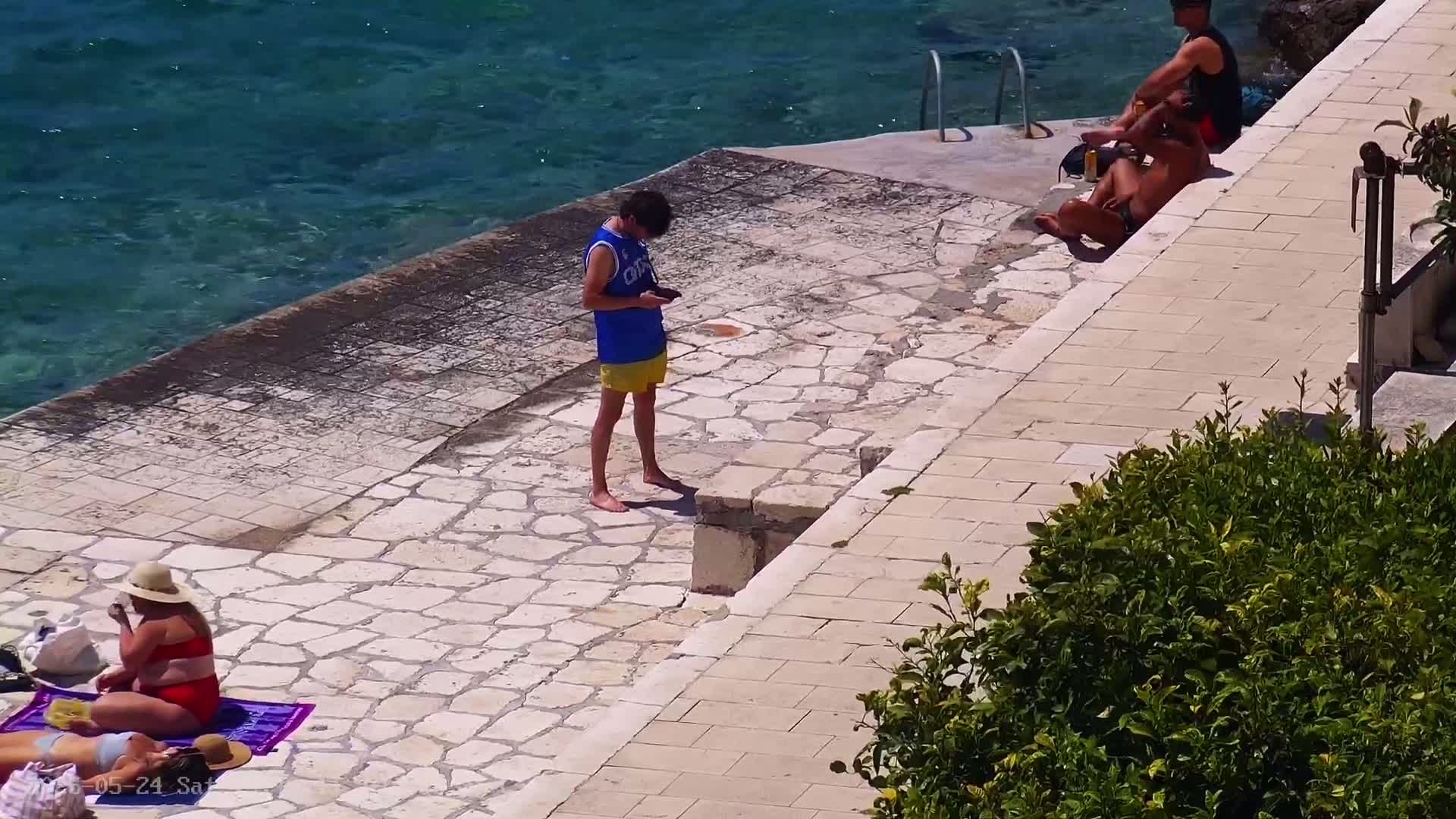 A stone patio next to clear blue water has several people relaxing and sunbathing on a sunny day.