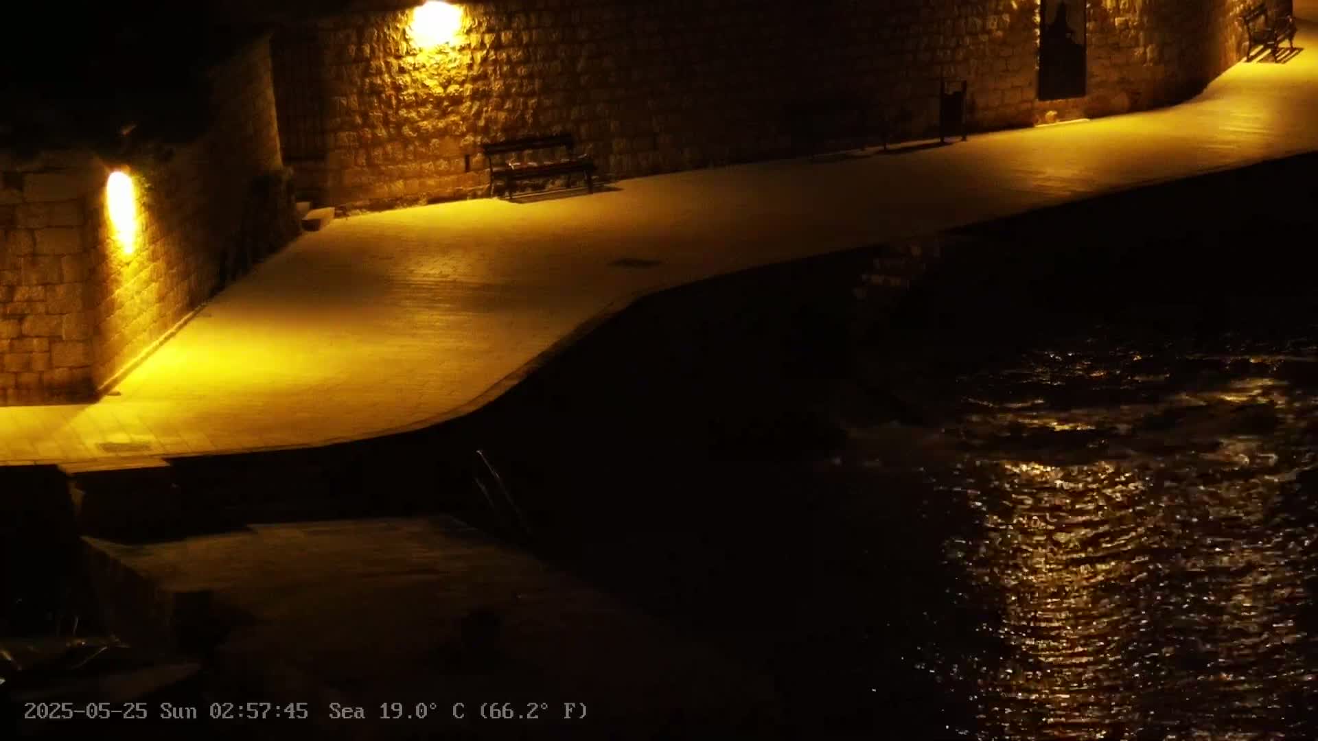 A nighttime, illuminated stone walkway curves alongside a dark body of water with gently rippling surfaces reflecting light.