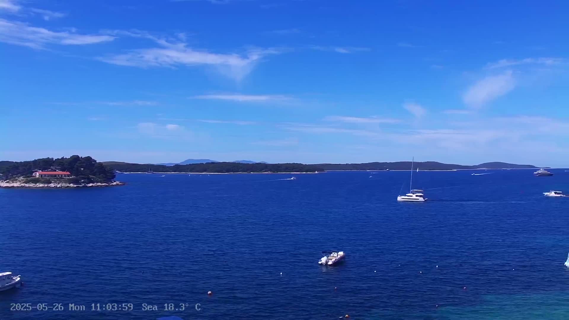 A tranquil blue sea scene under a mostly clear sky shows a small island with a building, several boats, and a distant shoreline.