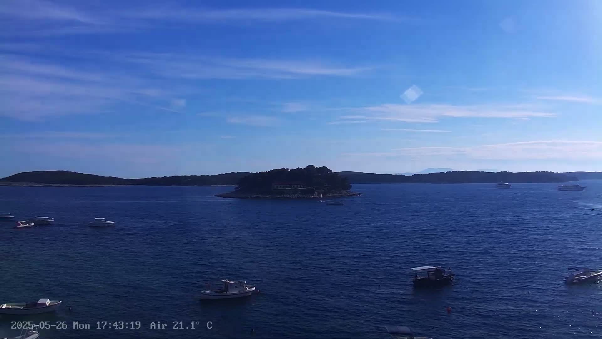 A small island with buildings sits in a calm, blue sea under a mostly clear, bright blue sky, surrounded by several small boats.