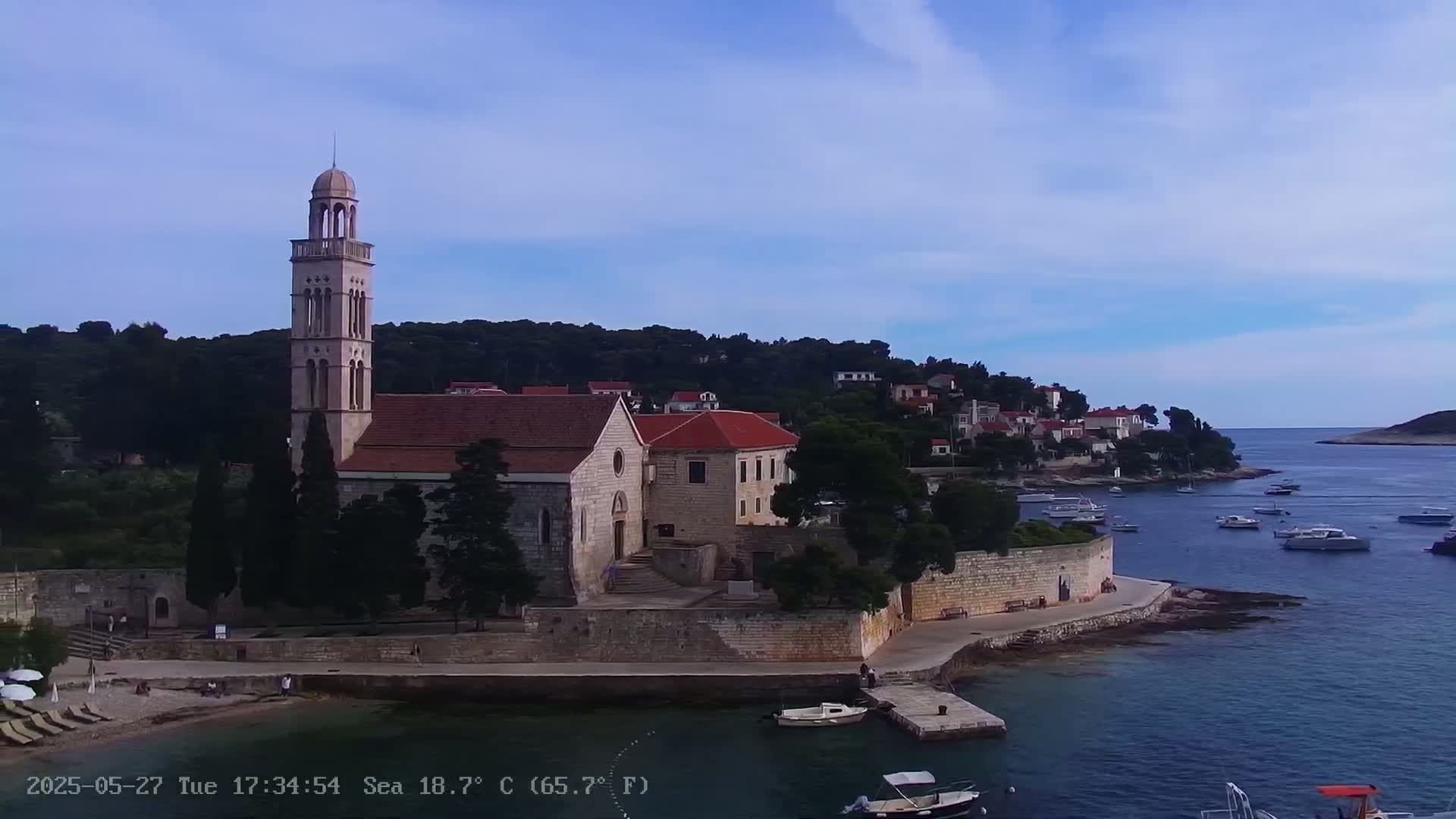 A church with a tall bell tower sits on a stone waterfront overlooking a calm bay with several boats under a mostly clear sky.
