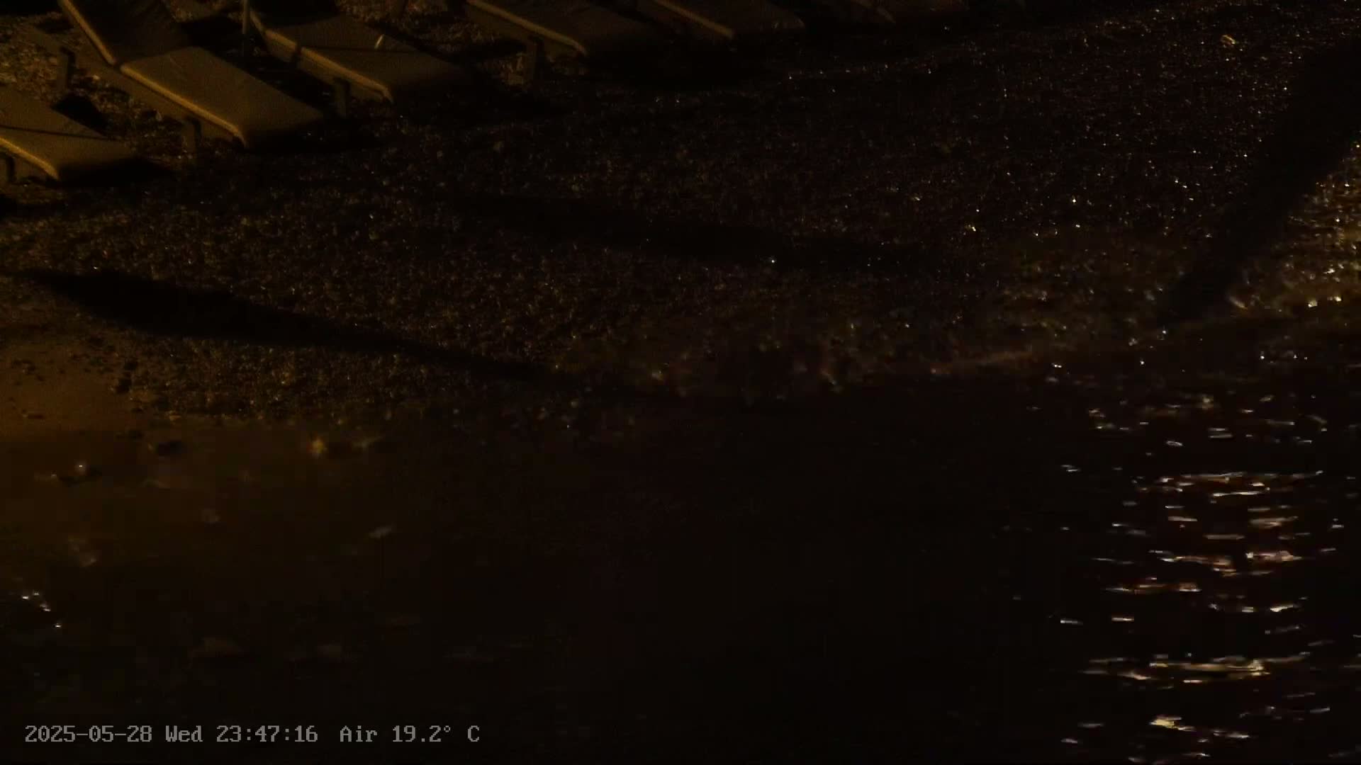 Dark brown lounge chairs cast shadows on a dark, pebbled beach at night next to gently lapping water.