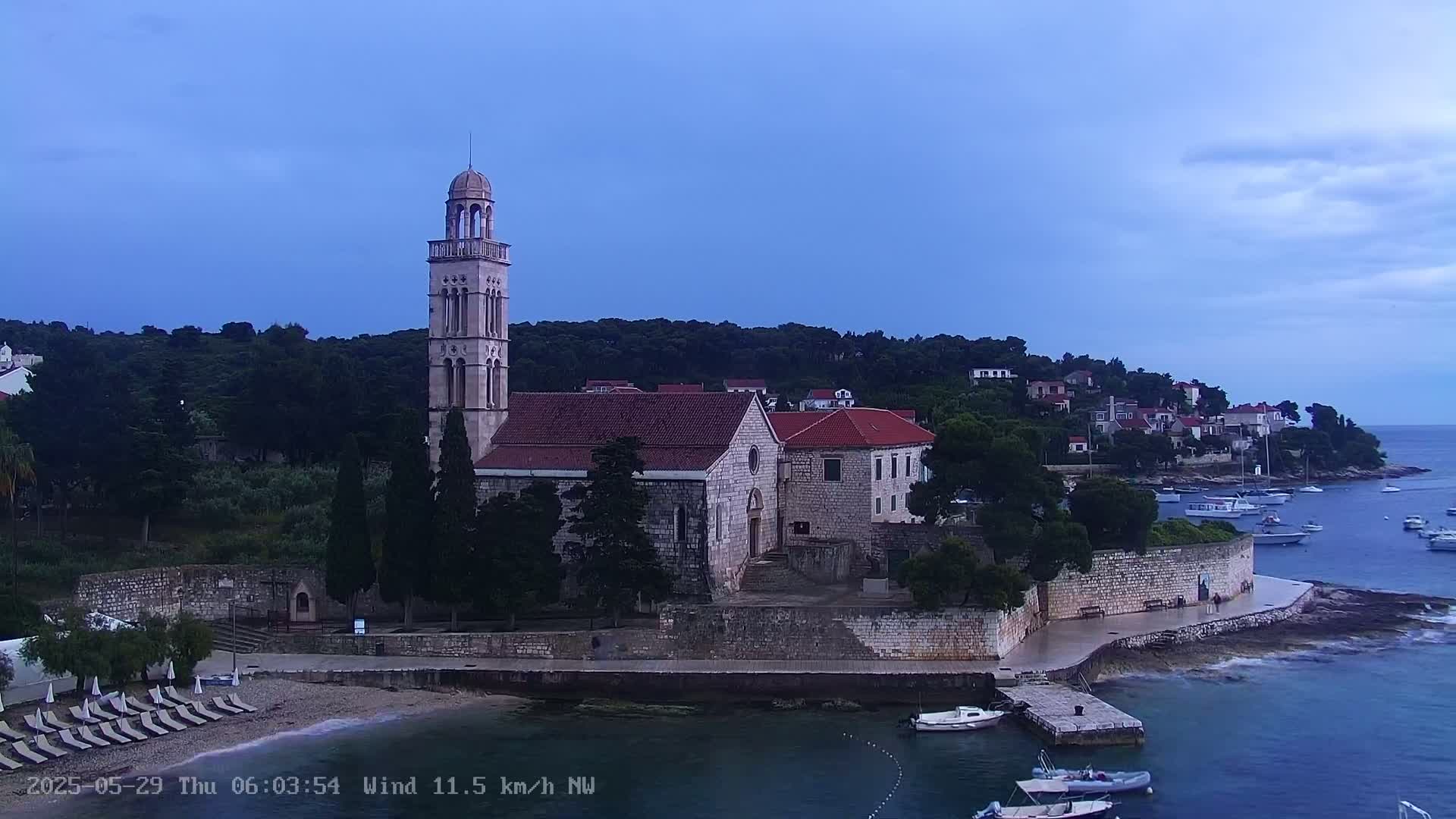 A stone church with a tall bell tower sits on a coastal hillside overlooking a calm bay with several boats under a twilight sky.