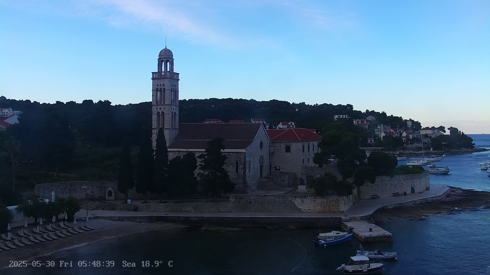 A stone church with a tall bell tower sits on a waterfront, next to a calm sea under a clear, twilight sky.