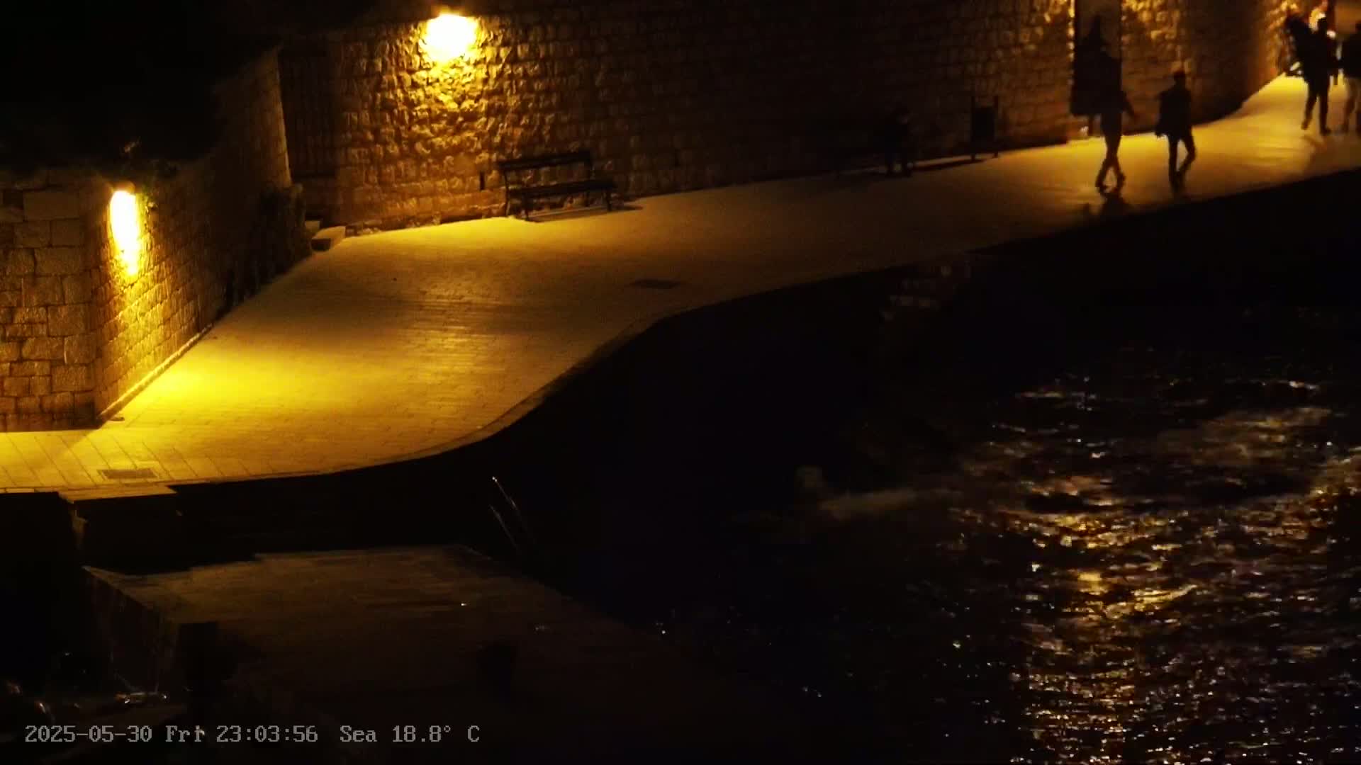 A stone walkway lit by lamps at night curves gently toward a dark body of water with small waves reflecting light.