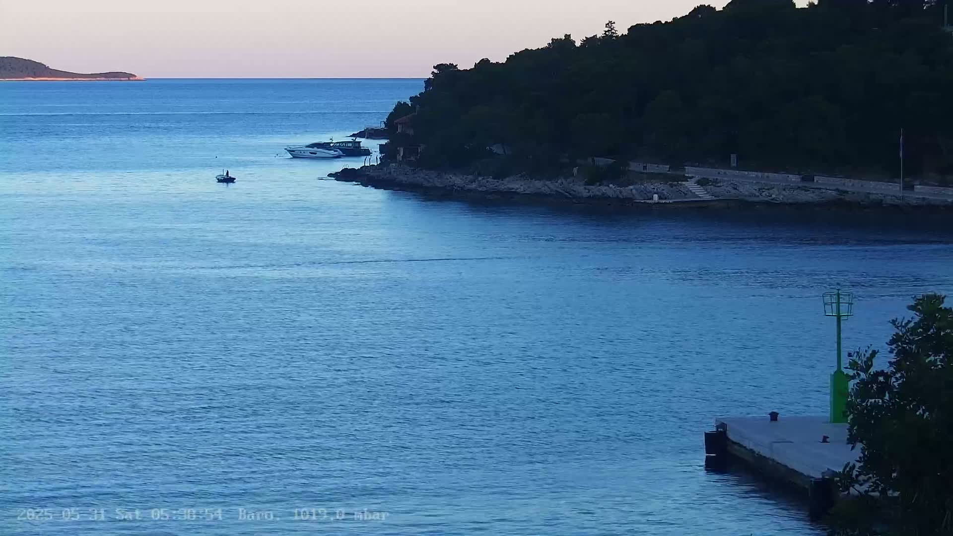 A calm, blue sea at dusk, with a small island in the distance, several boats, and a rocky shoreline with dark green vegetation.