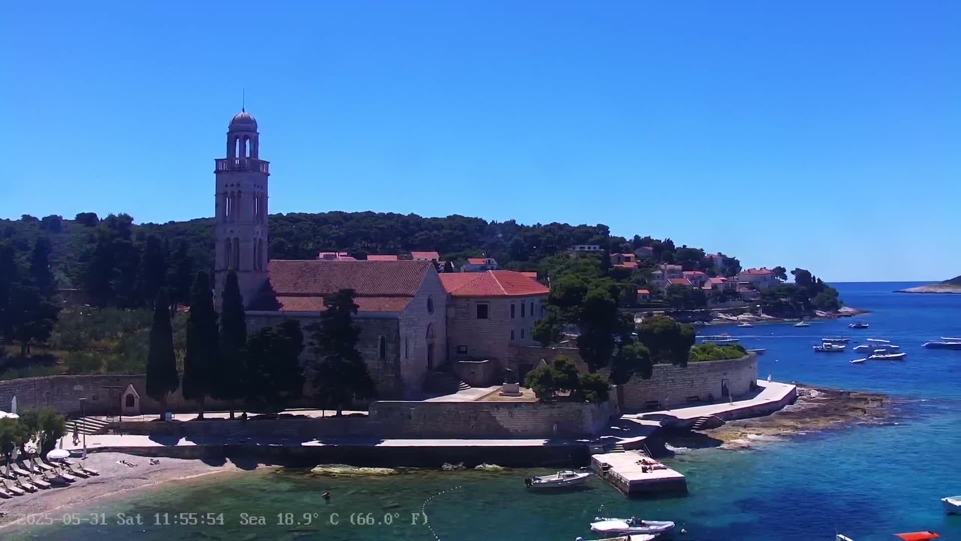 An aerial view shows a church with a tall bell tower situated on a rocky peninsula beside a calm, clear blue sea on a sunny day.