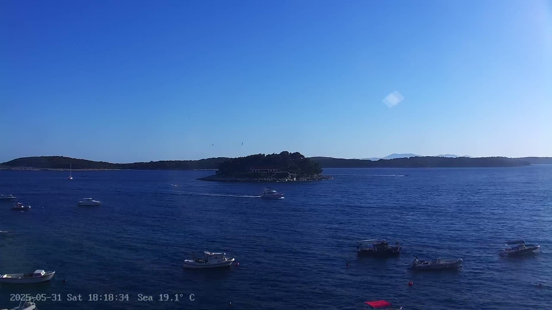Several small boats are scattered across a calm, dark blue sea, with a small, tree-covered island in the middle distance under a clear, bright blue sky.