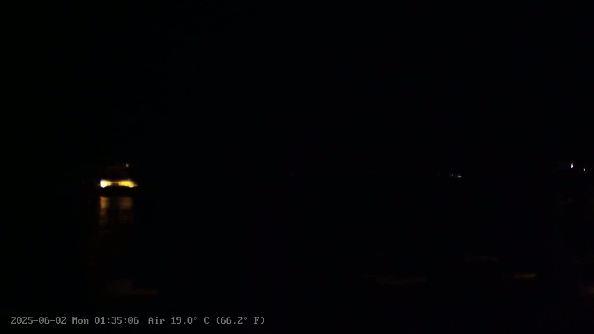 A dark nighttime scene shows a faintly illuminated structure reflected in dark water, with other distant lights visible in the blackness.