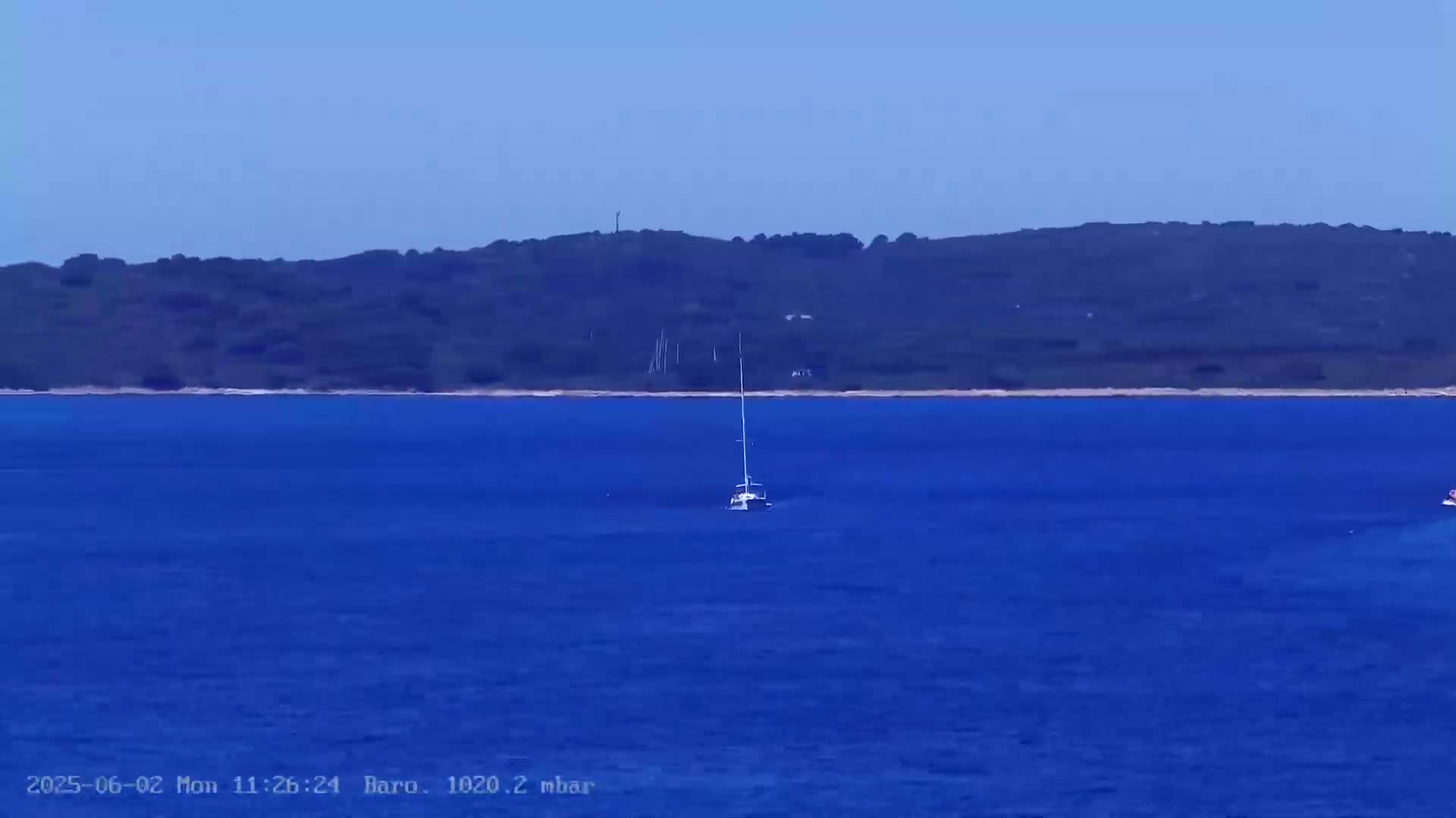 A sailboat sits on a calm, deep blue sea in front of a dark green, tree-covered coastline under a clear blue sky.