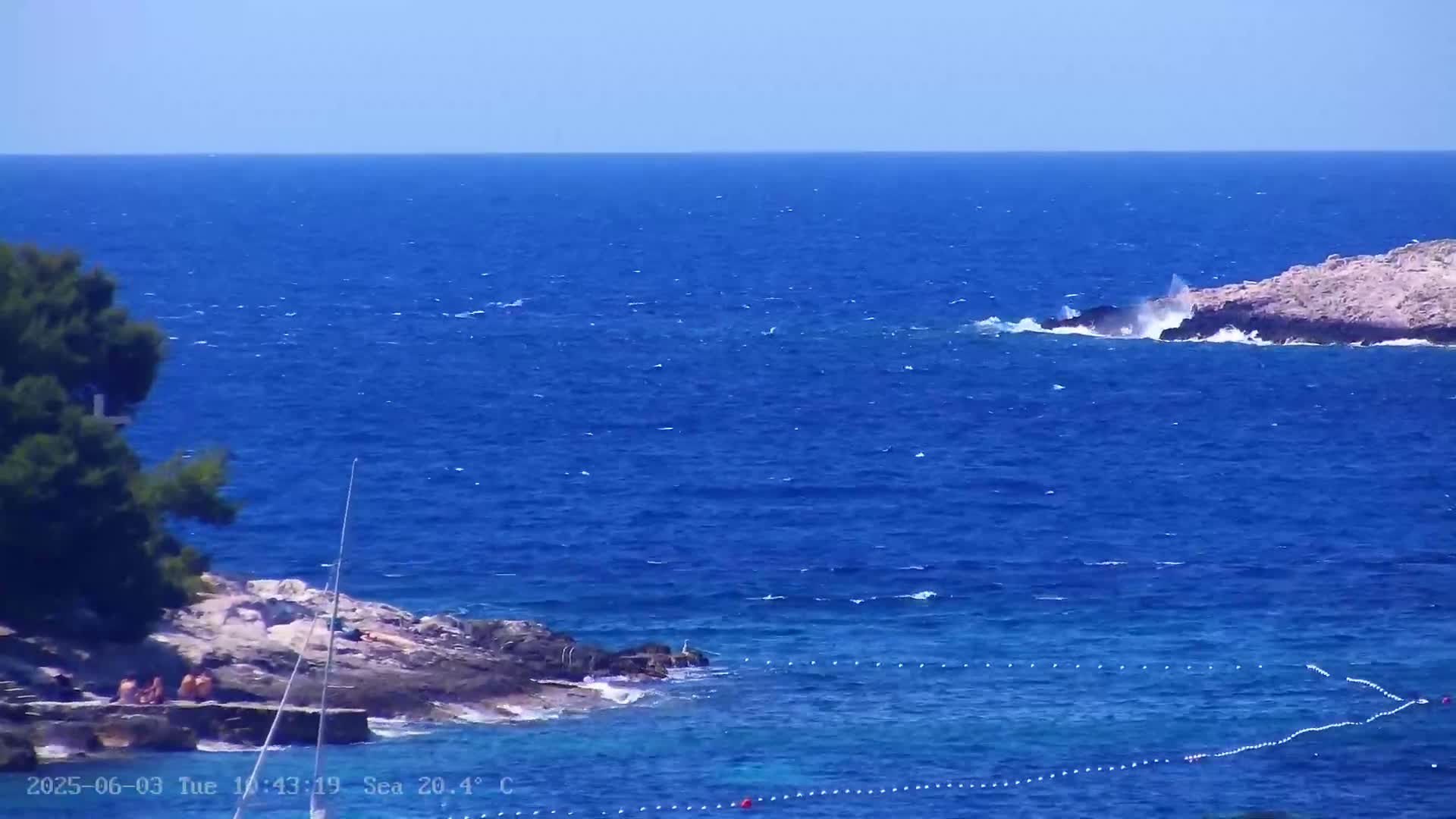A rocky shoreline with people sunbathing is partially visible beside a deep blue sea with small waves under a clear sky.