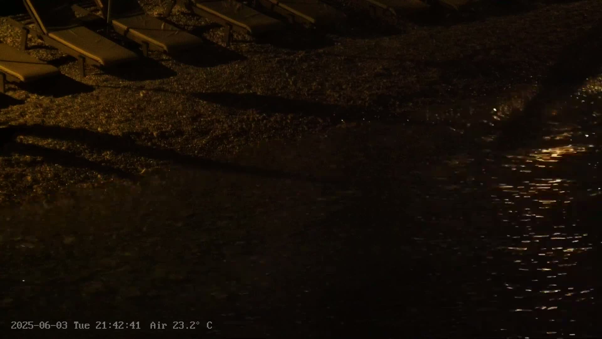 Several empty lounge chairs sit on a pebble beach at night next to dark water reflecting light.