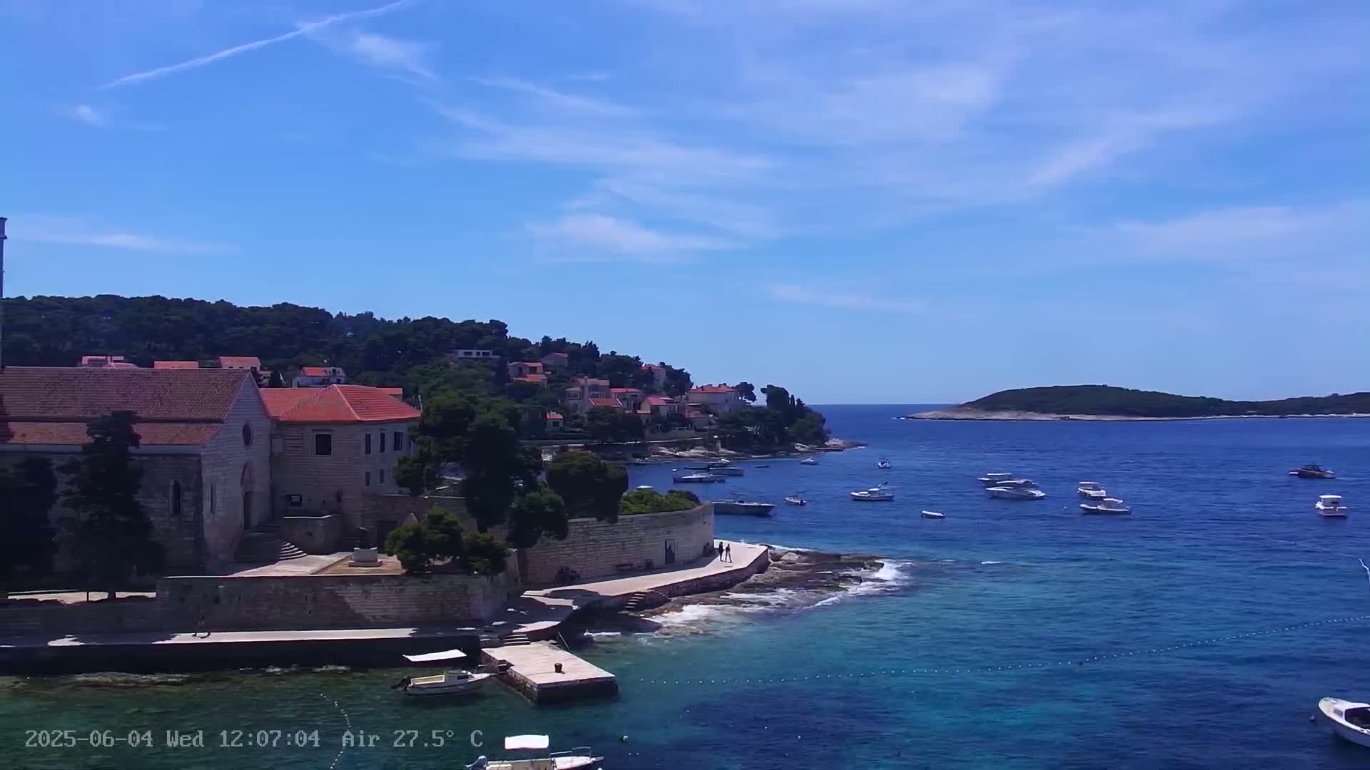 A stone church and buildings sit on a rocky coast overlooking a calm, blue sea dotted with small boats under a clear, sunny sky.