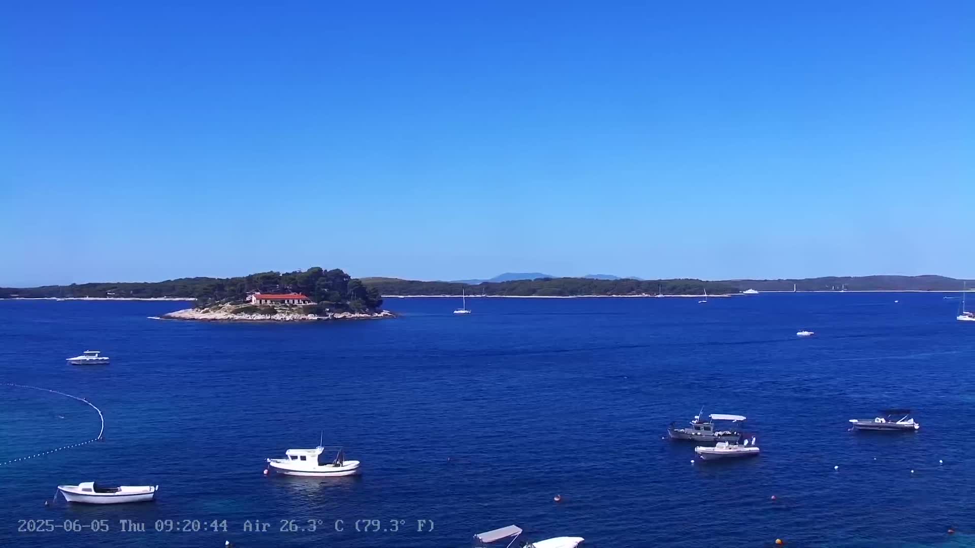 A small island with a building is visible in a calm, clear blue sea under a bright sunny sky, with several boats anchored nearby.