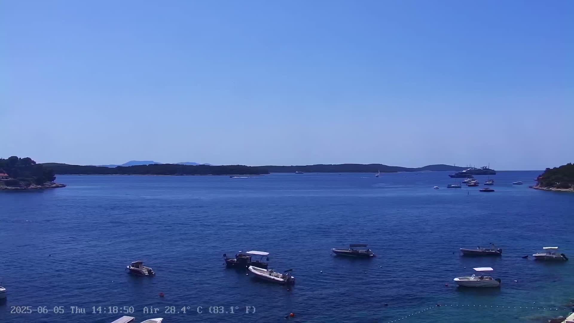 Several small boats are anchored in a calm, blue bay under a clear sky, with larger boats and a distant shoreline visible.