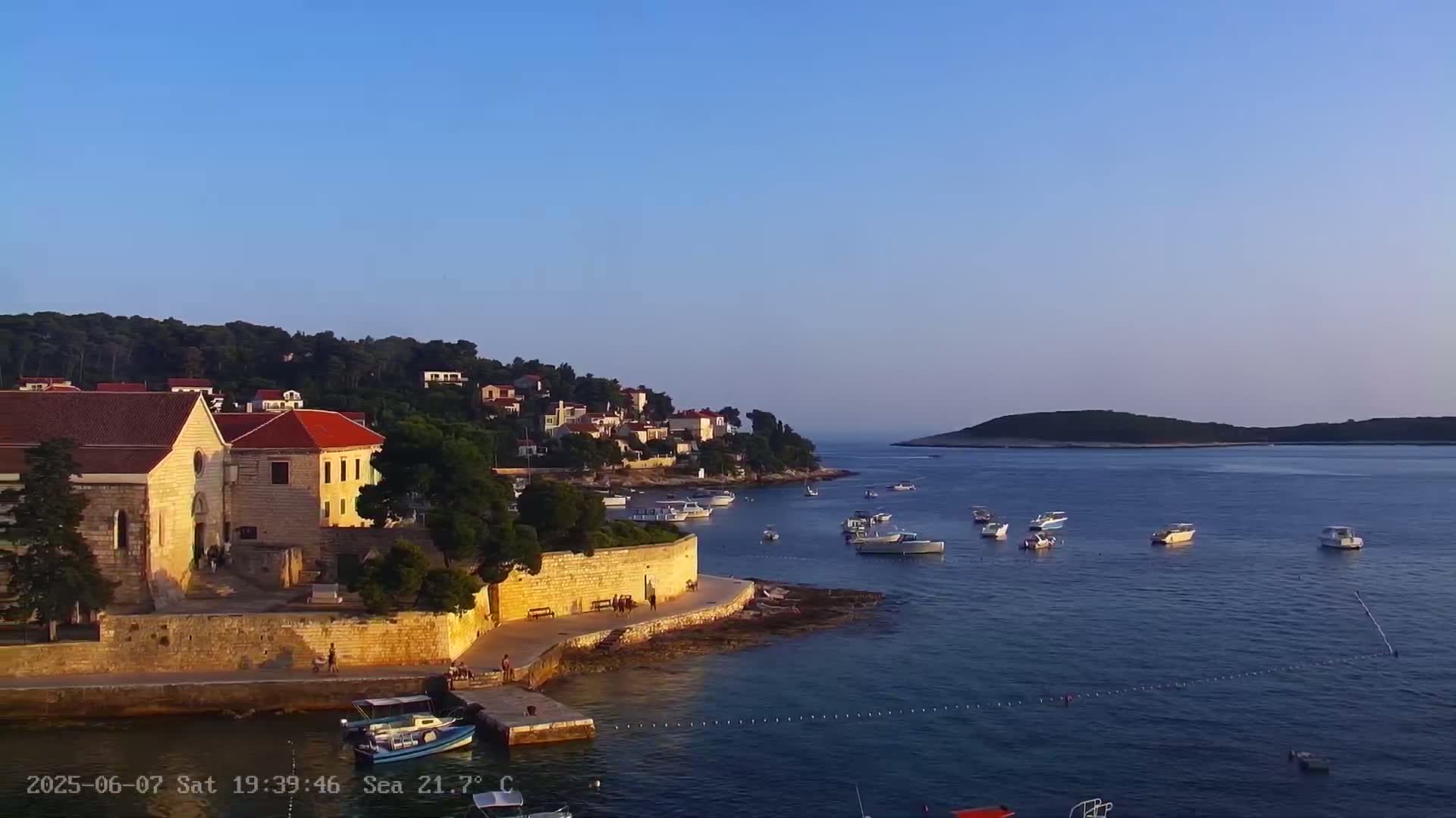 A stone building with red-tiled roofs sits on a calm sea under a clear blue sky, with several boats moored in the water and a small island visible in the distance.