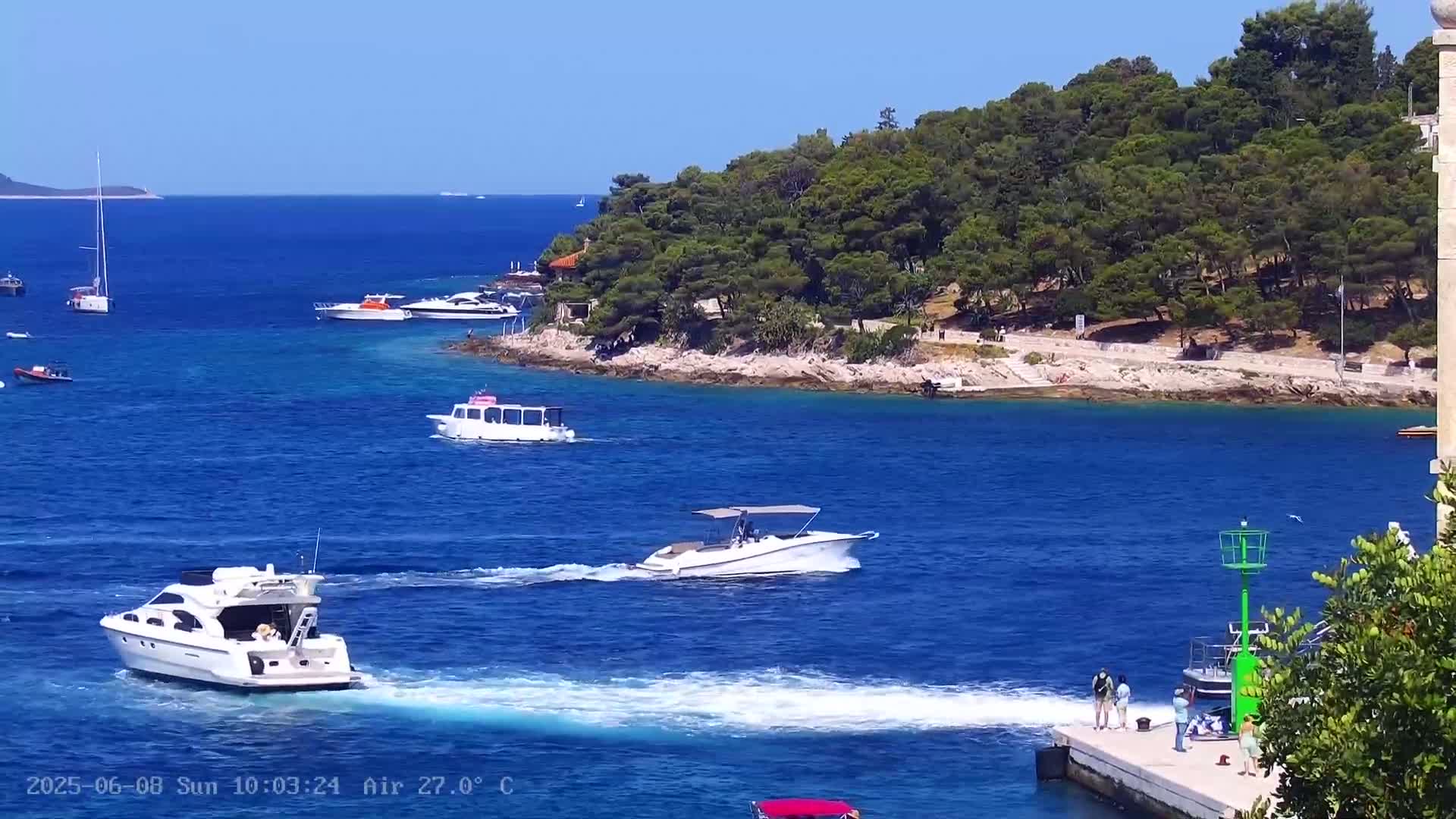 Several boats are in a clear blue body of water near a green, tree-covered island on a sunny day with an air temperature of 27°C.
