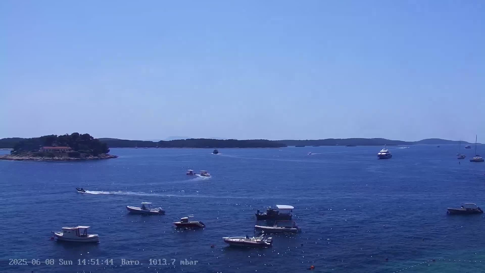 A calm, sunny day shows a variety of boats on a dark blue sea near a small island with buildings.
