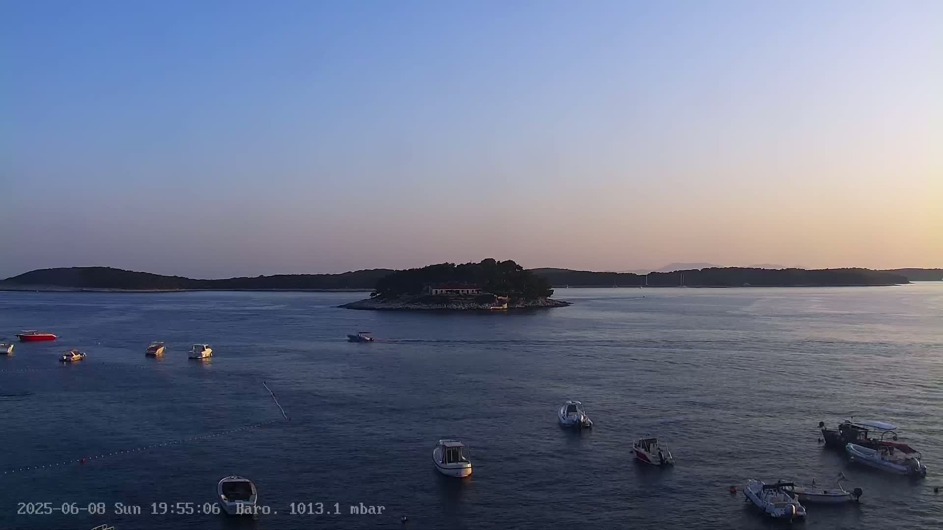 Several small boats are scattered across a calm body of water at sunset, with a small, tree-covered island in the mid-ground and larger landmasses in the background.