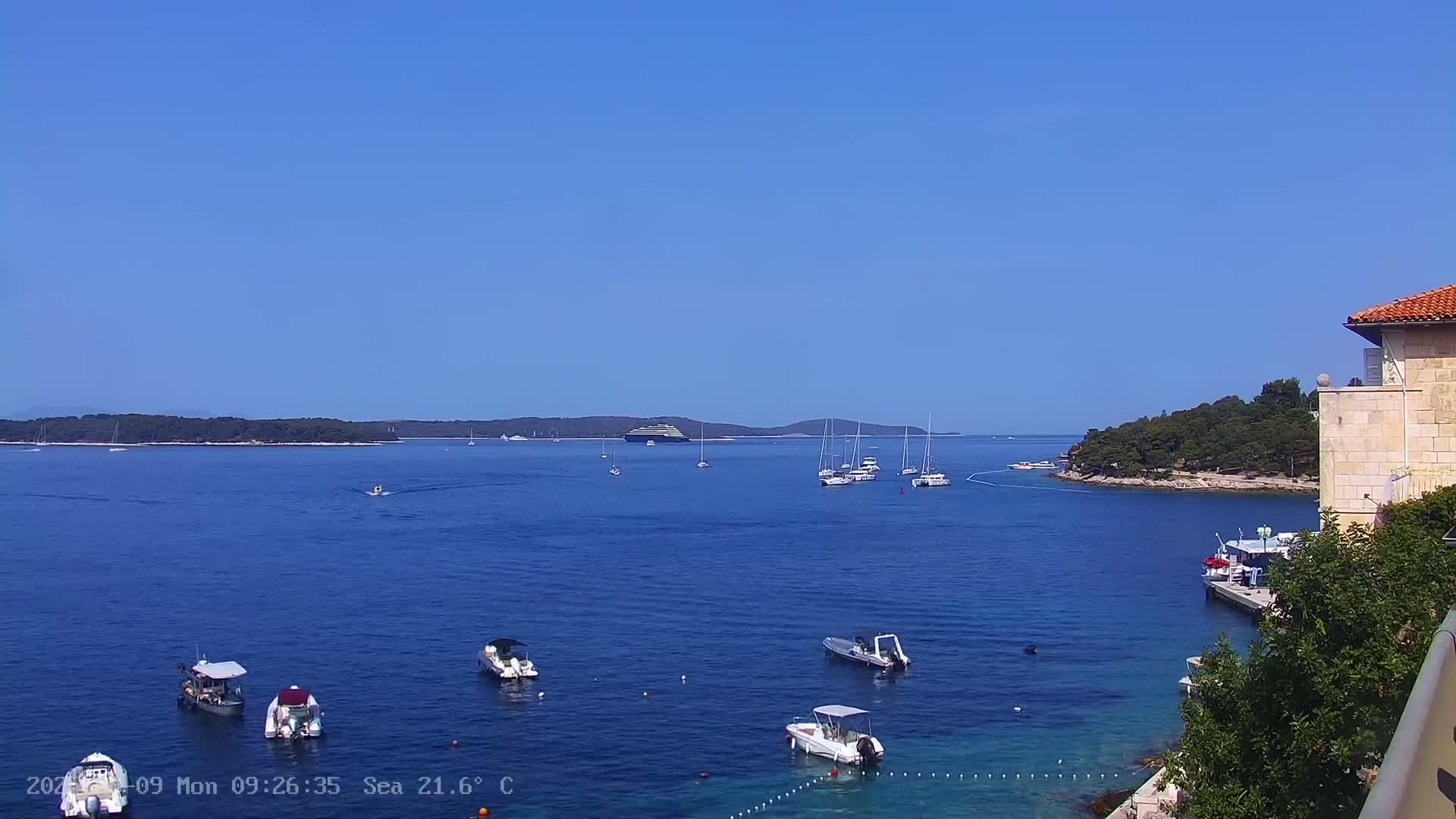 A clear, sunny day shows a calm blue sea with several boats and a large ship in the distance, near a small, tree-covered island and a stone building.