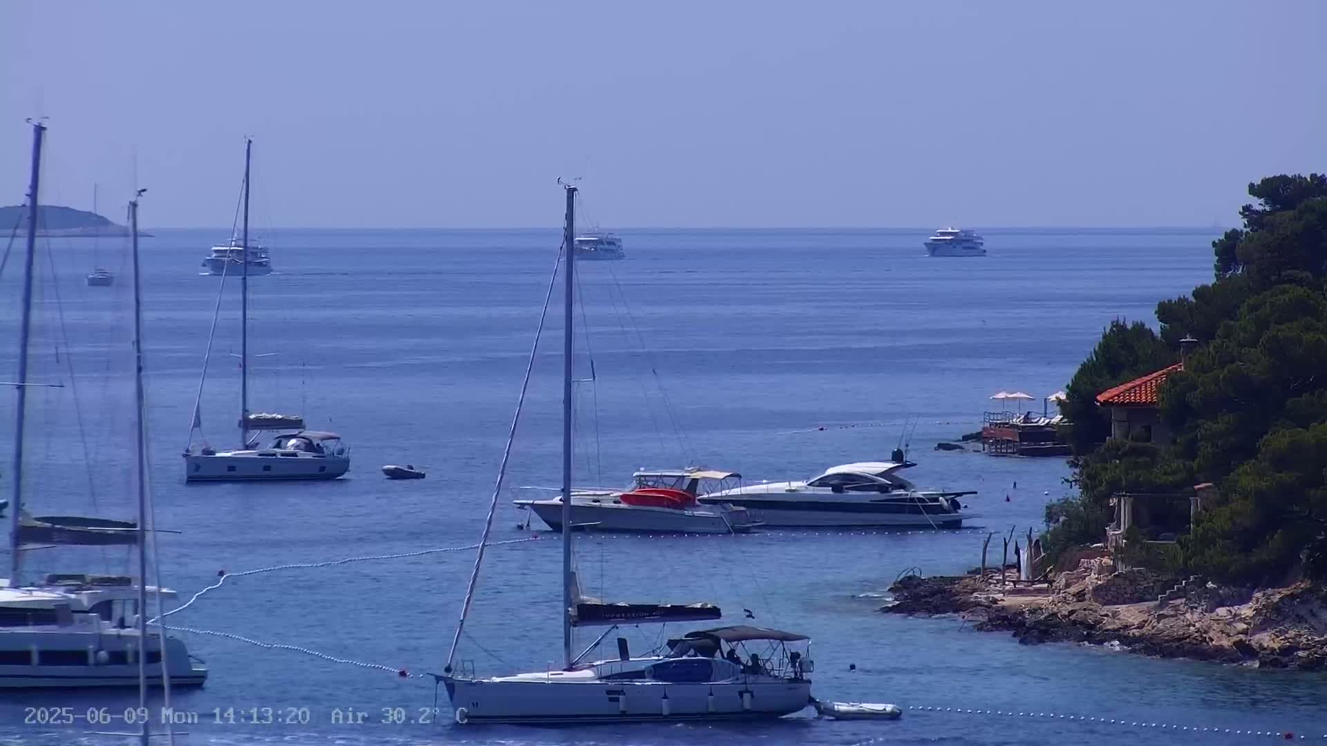 Several sailboats and motorboats are anchored in a calm, clear blue sea near a rocky shore with a small house and some trees on a sunny day.
