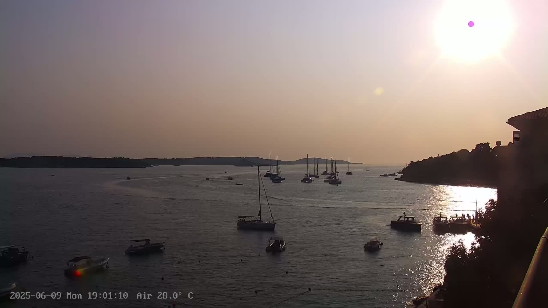 Many sailboats and motorboats are moored in a calm bay at sunset under a clear, warm sky.