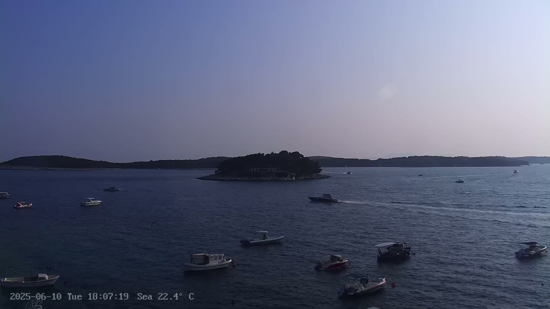 Several small boats are scattered across a calm body of water near a small, tree-covered island under a clear, light-blue sky.
