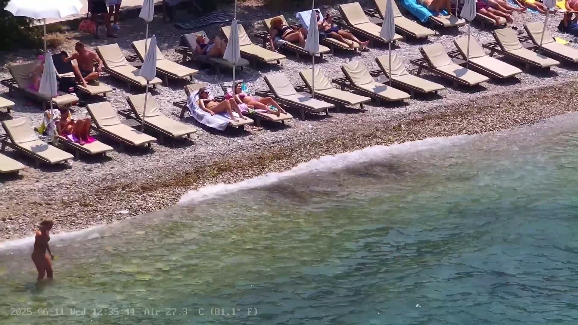 A pebble beach with numerous lounge chairs and beach umbrellas, occupied by sunbathers, borders a calm, clear, teal-colored body of water.