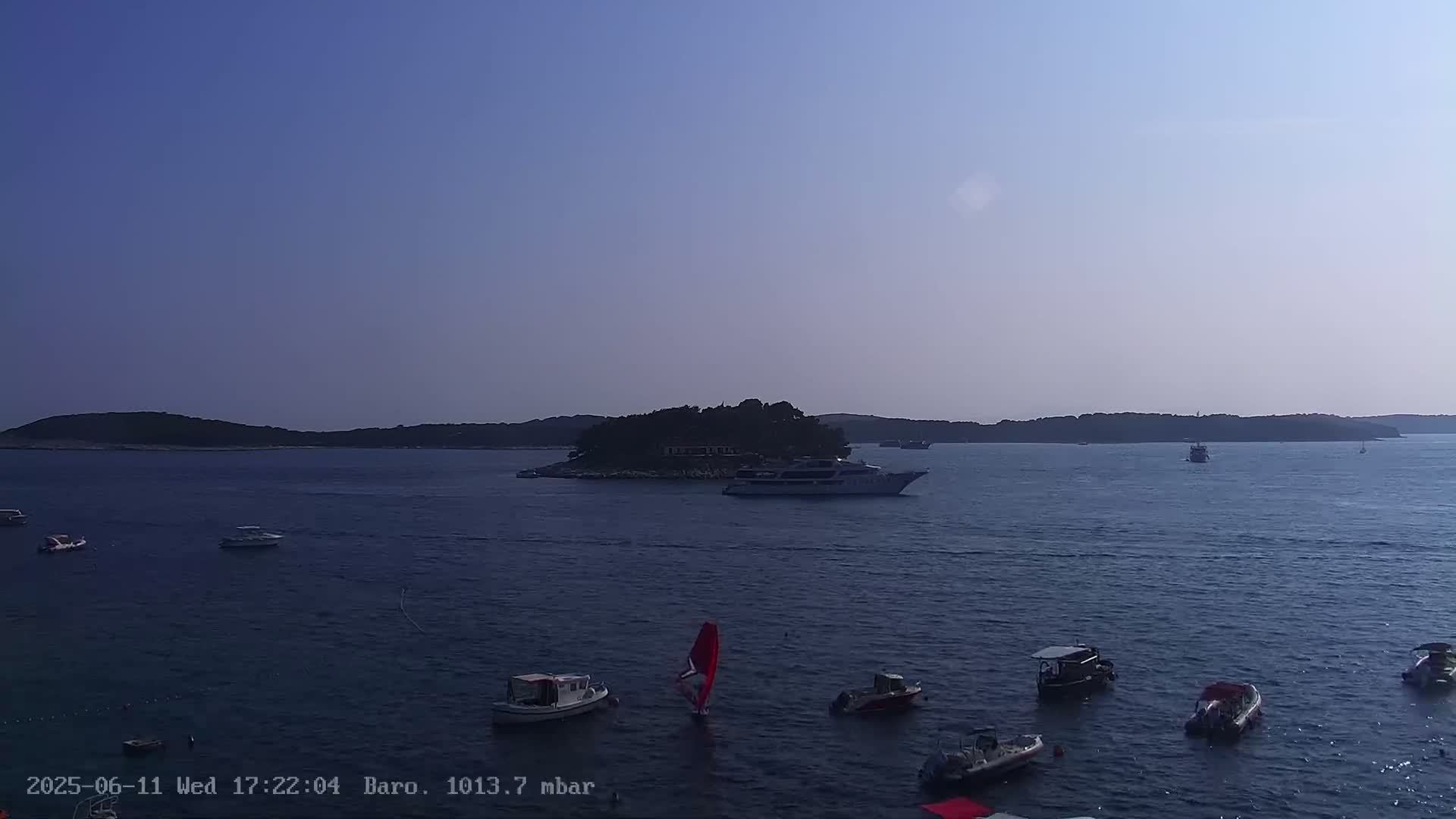A calm sea with numerous small boats and a larger vessel near a small, tree-covered island under a clear, light-blue sky.