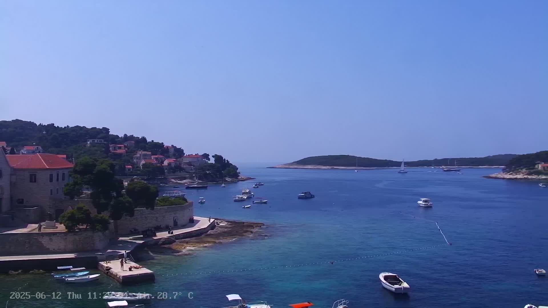 A clear, sunny day reveals a tranquil coastal scene with a village of red-roofed houses nestled on a hillside overlooking a calm, blue bay dotted with boats.