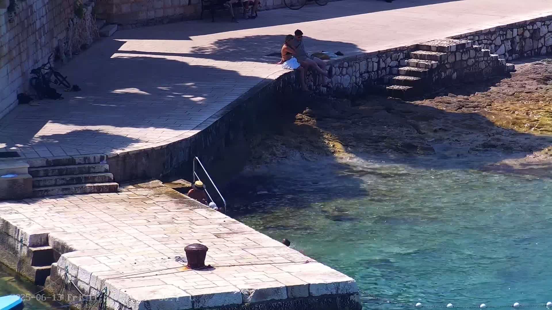 A stone dock with people sitting and entering clear, shallow water on a sunny day.