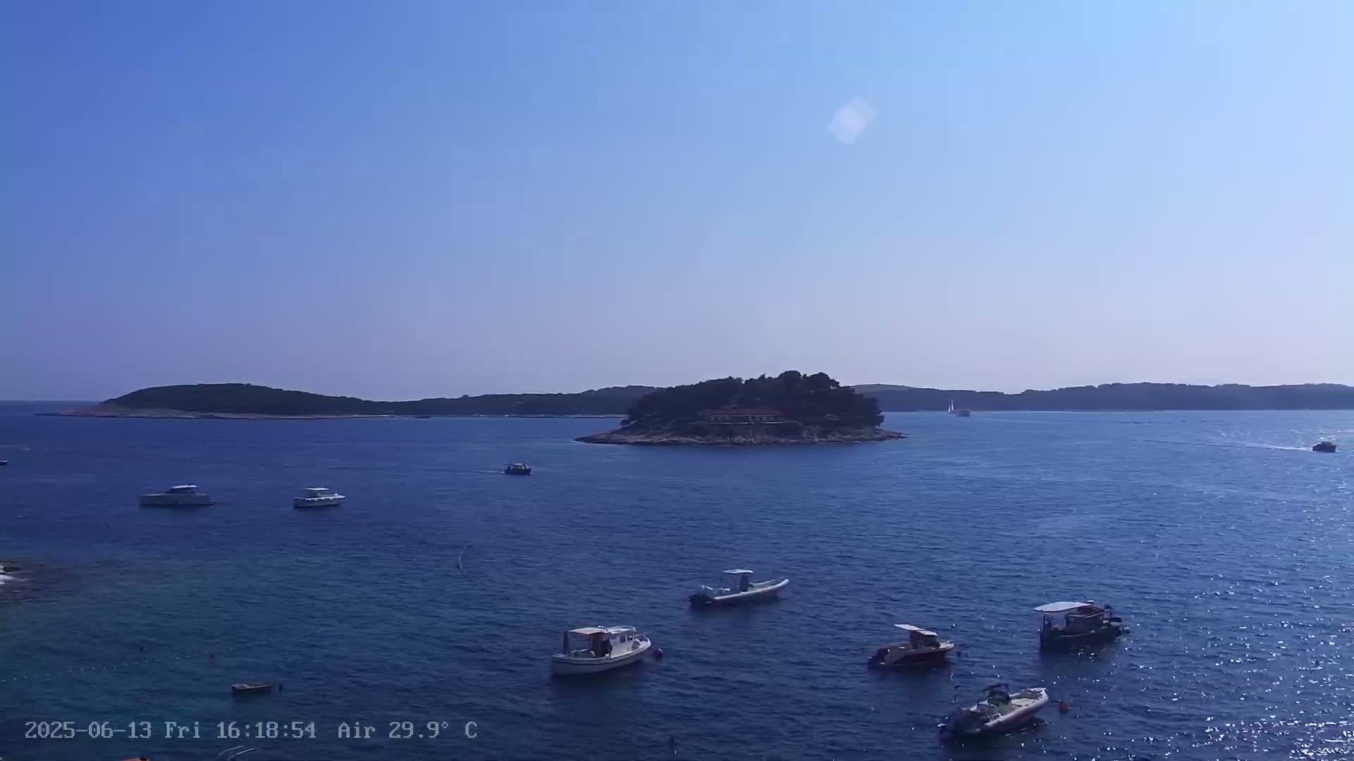Several small boats are scattered across a calm, blue sea, with a small, tree-covered island in the mid-ground under a clear, sunny sky.