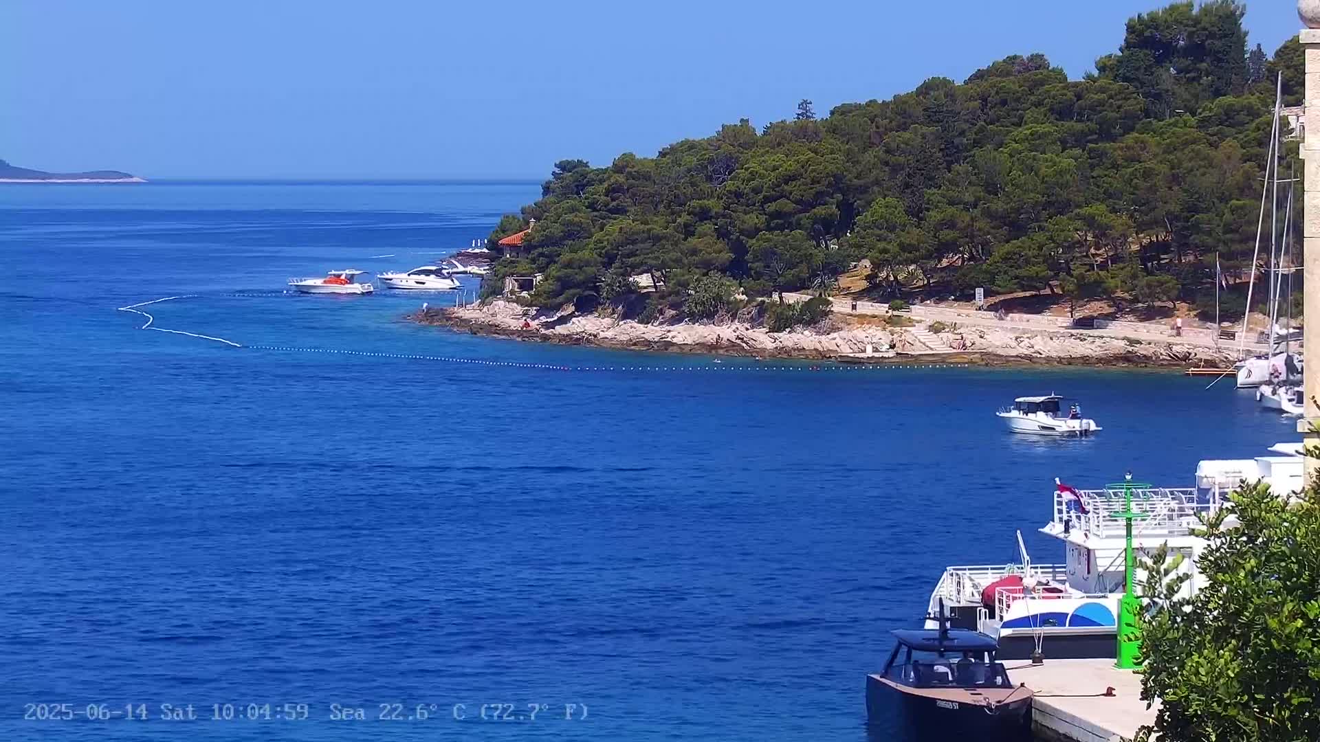 A picturesque, sunny waterfront scene shows several boats moored near a verdant, tree-covered island with a rocky shoreline.